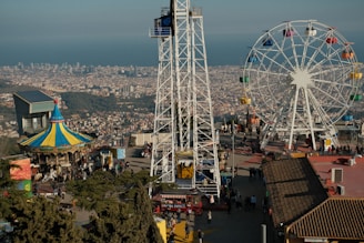 An amusement park scene with a large Ferris wheel featuring multi-colored gondolas, a brightly colored carousel with a pointed roof, and a panoramic view of a cityscape in the background. The foreground includes walkways with numerous people enjoying the attractions, and a range of structures and trees also populate the area.