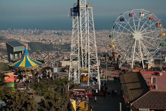 An amusement park scene with a large Ferris wheel featuring multi-colored gondolas, a brightly colored carousel with a pointed roof, and a panoramic view of a cityscape in the background. The foreground includes walkways with numerous people enjoying the attractions, and a range of structures and trees also populate the area.