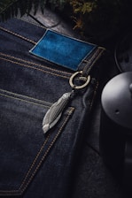 A close-up view of dark blue denim jeans with yellow stitching. The jeans have a blue leather patch and a metallic feather keychain attached to a ring near the pocket. Surrounding elements include fern leaves and a ceramic or metal object, possibly a mug or pot.