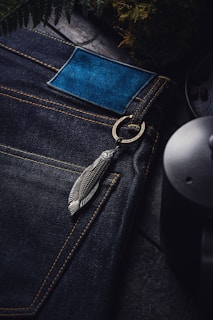 A close-up view of dark blue denim jeans with yellow stitching. The jeans have a blue leather patch and a metallic feather keychain attached to a ring near the pocket. Surrounding elements include fern leaves and a ceramic or metal object, possibly a mug or pot.