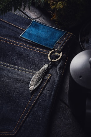 A close-up view of dark blue denim jeans with yellow stitching. The jeans have a blue leather patch and a metallic feather keychain attached to a ring near the pocket. Surrounding elements include fern leaves and a ceramic or metal object, possibly a mug or pot.