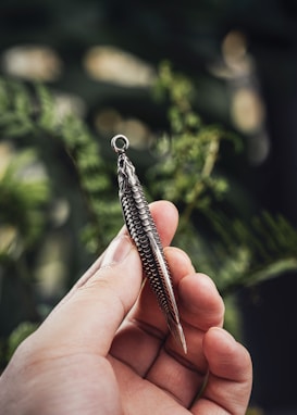 A hand is holding a metallic pendant shaped like a fish with intricate scale details. The background is softly blurred with hints of greenery, suggesting an outdoor setting.