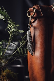 A close-up view of a brown leather bag with intricate detailing and a decorative keychain shaped like a fish hanging from a metal clasp. In the background, green fern leaves add a natural element, with a soft focus on a dark backdrop.