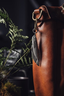 A close-up view of a brown leather bag with intricate detailing and a decorative keychain shaped like a fish hanging from a metal clasp. In the background, green fern leaves add a natural element, with a soft focus on a dark backdrop.