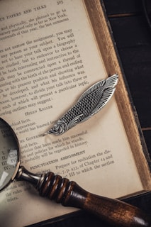 A vintage open book displaying text with a metal bookmark shaped like a fish resting on its pages. A magnifying glass with a wooden handle lies beside the book, partially over the text.