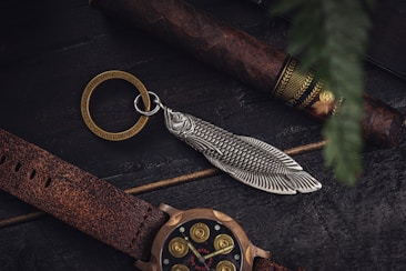 A rustic scene featuring a cigar with ornate bands, a keychain with a fish-shaped charm, and a watch with a leather strap lying on a dark wooden surface.