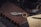 A vintage-style keychain with feather details lies on a brown leather-bound notebook. Nearby, there is a black coffee cup on a saucer surrounded by scattered coffee beans. A handwritten letter and a black pen complete the arrangement on a dark wooden surface.