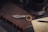 A vintage-style keychain with feather details lies on a brown leather-bound notebook. Nearby, there is a black coffee cup on a saucer surrounded by scattered coffee beans. A handwritten letter and a black pen complete the arrangement on a dark wooden surface.