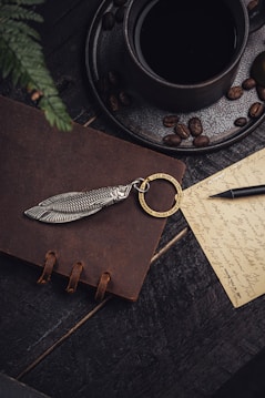 A dark-toned, moody composition features a black coffee cup on a saucer scattered with coffee beans, sitting on a wooden surface. A brown leather notebook adorned with a unique silver fish-shaped keychain and a gold ring engraved with 'Get busy living or get busy dying' is nearby. A handwritten note with cursive writing and a black fountain pen add a touch of vintage elegance.