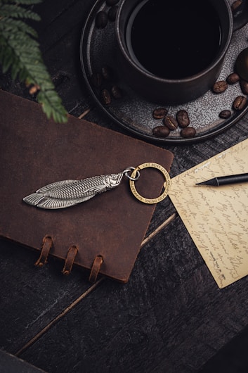 A dark-toned, moody composition features a black coffee cup on a saucer scattered with coffee beans, sitting on a wooden surface. A brown leather notebook adorned with a unique silver fish-shaped keychain and a gold ring engraved with 'Get busy living or get busy dying' is nearby. A handwritten note with cursive writing and a black fountain pen add a touch of vintage elegance.