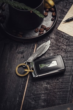 A detailed car key fob with a decorative feather-shaped keychain attached is positioned on a rustic wooden surface. Coffee beans are scattered nearby, with a dark, textured coffee cup and saucer in the background. A handwritten note and pen add to the cozy, vintage atmosphere.