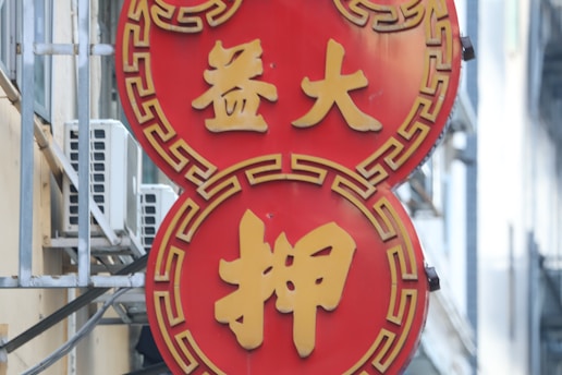 A vibrant red and gold sign with Chinese characters is mounted on the side of a building. The sign features a double circular design with geometric patterns around the edges.