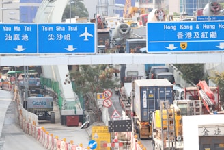 A busy urban scene with traffic congestion, featuring a variety of vehicles such as trucks and buses. The overhead road signs indicate directions to places like Yau Ma Tei and Tsim Sha Tsui, as well as the Hong Kong & Hung Hom area. Construction equipment and orange and white barricades line the roadway, suggesting ongoing roadwork. A few trees are visible alongside the road.