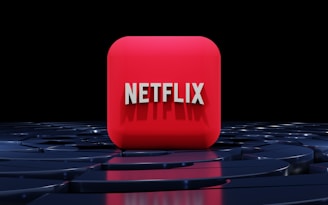 A red, cube-shaped icon with the word 'NETFLIX' prominently displayed in white, bold letters. The cube is set against a dark, glossy, circular patterned background, reflecting a sleek, futuristic design.