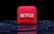 A red, cube-shaped icon with the word 'NETFLIX' prominently displayed in white, bold letters. The cube is set against a dark, glossy, circular patterned background, reflecting a sleek, futuristic design.