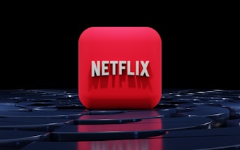 A red, cube-shaped icon with the word 'NETFLIX' prominently displayed in white, bold letters. The cube is set against a dark, glossy, circular patterned background, reflecting a sleek, futuristic design.