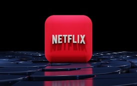 A red, cube-shaped icon with the word 'NETFLIX' prominently displayed in white, bold letters. The cube is set against a dark, glossy, circular patterned background, reflecting a sleek, futuristic design.