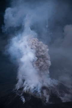 A large plume of ash and smoke billows from the crater of an erupting volcano. The surrounding area is dark and shrouded in shadow, highlighting the intensity of the eruption. Thick smoke rises vertically into the sky, with varying shades of gray creating a dramatic contrast against the darker landscape.