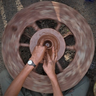 Hands are shaping a clay pot on a spinning pottery wheel. The image captures the motion blur of the wheel, while the person's hands are focused on molding the clay. The setting appears earthy and natural.