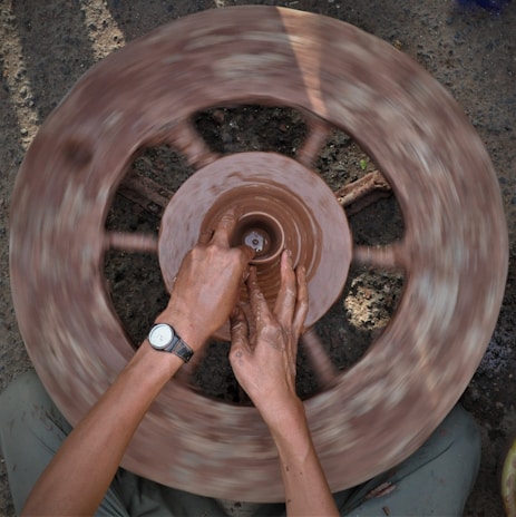 Hands are shaping a clay pot on a spinning pottery wheel. The image captures the motion blur of the wheel, while the person's hands are focused on molding the clay. The setting appears earthy and natural.