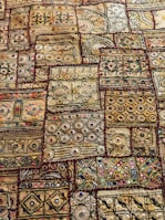 An intricate textile displaying a variety of geometric patterns and shapes, including circles, squares, and lines. The fabric features a rich tapestry of embroidery with numerous colors and textures, showcasing a traditional or cultural aesthetic. The designs are symmetrically arranged in patches, each with unique motifs and vibrant detailing.