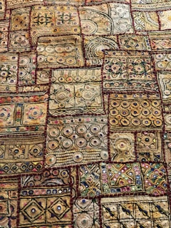 An intricate textile displaying a variety of geometric patterns and shapes, including circles, squares, and lines. The fabric features a rich tapestry of embroidery with numerous colors and textures, showcasing a traditional or cultural aesthetic. The designs are symmetrically arranged in patches, each with unique motifs and vibrant detailing.