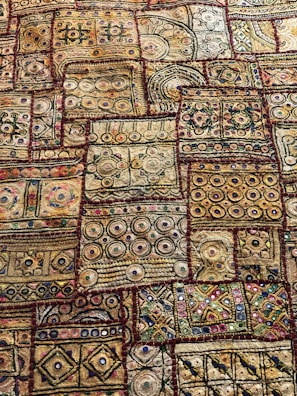An intricate textile displaying a variety of geometric patterns and shapes, including circles, squares, and lines. The fabric features a rich tapestry of embroidery with numerous colors and textures, showcasing a traditional or cultural aesthetic. The designs are symmetrically arranged in patches, each with unique motifs and vibrant detailing.