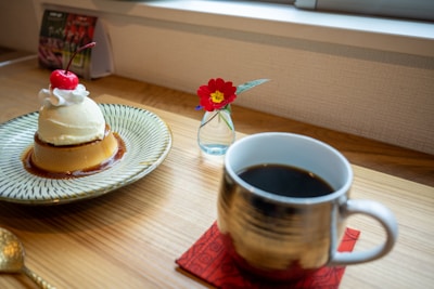 A close-up view of a dessert table setup includes a caramel pudding topped with vanilla ice cream, whipped cream, and a cherry on a decorative plate. A stylish metal mug filled with black coffee is placed on a red coaster nearby. A small vase with a single bright red flower adds a touch of color to the wooden table setting.
