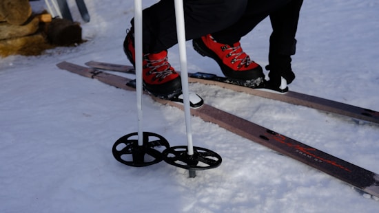 A close-up view of a person wearing red ski boots fastened onto wooden skis on a snowy surface. The individual is using ski poles, which have large circular baskets at their base. The snow is compacted, indicating a prepared ski area.