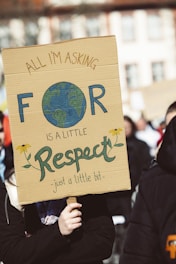 A person holds a cardboard protest sign with a message asking for respect. The sign features a hand-drawn Earth and flowers.