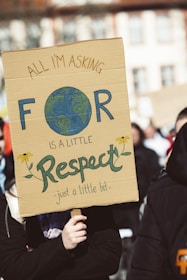 A person holds a cardboard protest sign with a message asking for respect. The sign features a hand-drawn Earth and flowers.