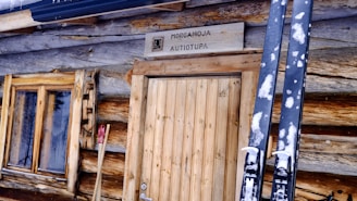 A rustic wooden cabin with a natural log construction features a wooden door and window. Skis are propped up against the cabin's exterior and a sign above the door displays the name 'Morgamoja Autiotupa,' indicating a wilderness hut or shelter.