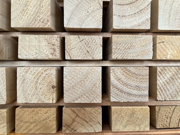 Several stacked wooden beams arranged in a neat, uniform pattern with visible grain and rings. The beams are light brown with textured surfaces.