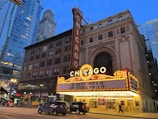 An iconic theater marquee in a city setting displays bright lights and signage with upcoming events. Skyscrapers and city architecture provide an urban backdrop as cars and pedestrians move along the street below.