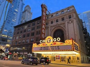 An iconic theater marquee in a city setting displays bright lights and signage with upcoming events. Skyscrapers and city architecture provide an urban backdrop as cars and pedestrians move along the street below.