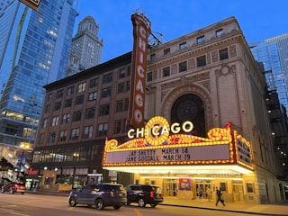 An iconic theater marquee in a city setting displays bright lights and signage with upcoming events. Skyscrapers and city architecture provide an urban backdrop as cars and pedestrians move along the street below.