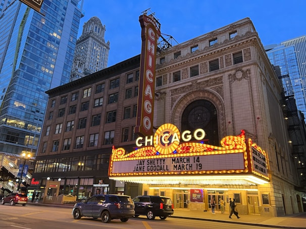An iconic theater marquee in a city setting displays bright lights and signage with upcoming events. Skyscrapers and city architecture provide an urban backdrop as cars and pedestrians move along the street below.