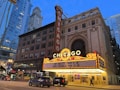 An iconic theater marquee in a city setting displays bright lights and signage with upcoming events. Skyscrapers and city architecture provide an urban backdrop as cars and pedestrians move along the street below.