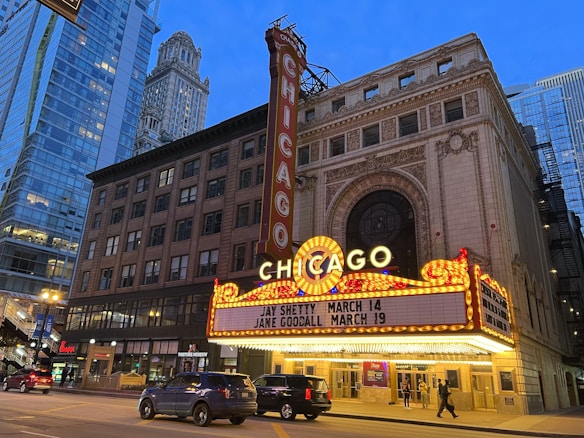An iconic theater marquee in a city setting displays bright lights and signage with upcoming events. Skyscrapers and city architecture provide an urban backdrop as cars and pedestrians move along the street below.