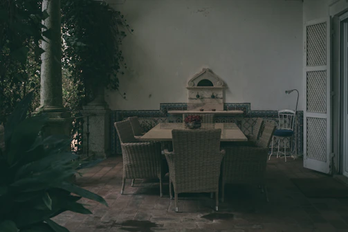 An outdoor patio area with a large wooden table surrounded by wicker chairs. The backdrop features a decorative stone wall with elegant tile work. Lush plants are visible on the left, adding a sense of privacy and tranquility to the space. Adjacent to the table is a smaller metal chair, next to a doorway with white, half-open shutters.