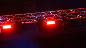 A metal truss structure is equipped with several stage lights. The stage lights are emitting a red glow, creating an intense and dramatic effect. The background is dark, highlighting the brightness and color of the lights.