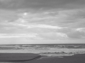 A grayscale photograph of an ocean scene with waves crashing onto the shore. The sky is overcast with a blanket of clouds, creating a dramatic and moody atmosphere. The shoreline appears at the bottom of the image, with the waves forming white crests as they approach the sand.