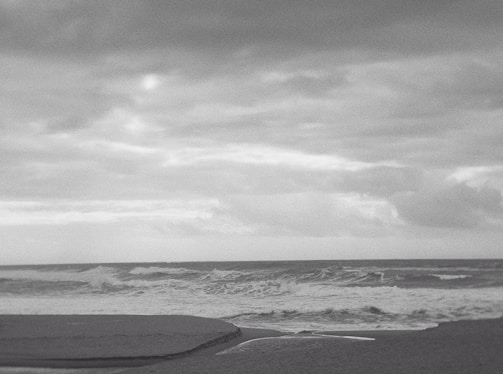 A grayscale photograph of an ocean scene with waves crashing onto the shore. The sky is overcast with a blanket of clouds, creating a dramatic and moody atmosphere. The shoreline appears at the bottom of the image, with the waves forming white crests as they approach the sand.