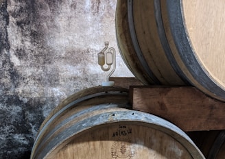 Several wooden barrels are stacked against a rustic, textured wall. One of the barrels has a small device attached to it, which appears to be a fermentation airlock.