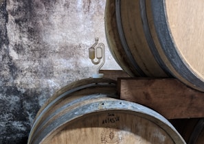 Several wooden barrels are stacked against a rustic, textured wall. One of the barrels has a small device attached to it, which appears to be a fermentation airlock.