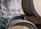 Several wooden barrels are stacked against a rustic, textured wall. One of the barrels has a small device attached to it, which appears to be a fermentation airlock.