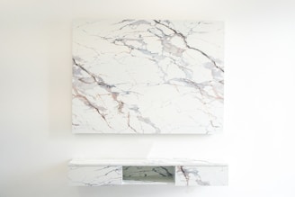 A room featuring a minimalist design with a large marble-patterned panel mounted on the wall. Below, a matching marble-patterned console table with open shelving appears attached to the wall. The decor is sleek and modern, emphasizing clean lines and a light color palette.