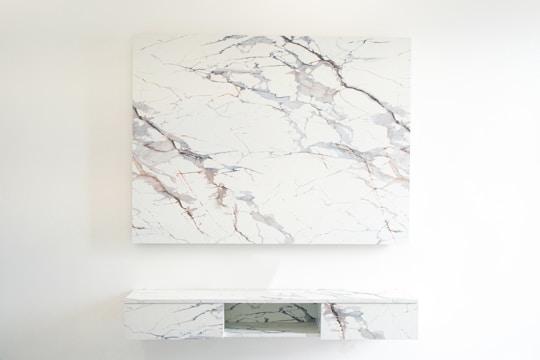 A room featuring a minimalist design with a large marble-patterned panel mounted on the wall. Below, a matching marble-patterned console table with open shelving appears attached to the wall. The decor is sleek and modern, emphasizing clean lines and a light color palette.