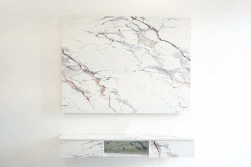 A room featuring a minimalist design with a large marble-patterned panel mounted on the wall. Below, a matching marble-patterned console table with open shelving appears attached to the wall. The decor is sleek and modern, emphasizing clean lines and a light color palette.