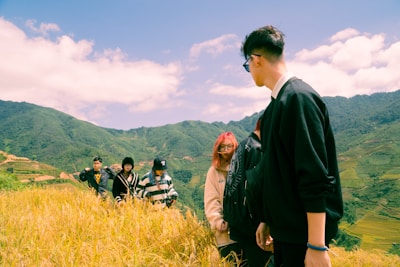 A group of young people stands on a hillside with rice fields in the foreground, surrounded by lush, green mountains under a partly cloudy sky. They are dressed in casual, warm clothing and appear to be enjoying the natural scenery.